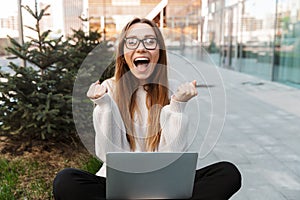 Beautiful excited shocked young happy business woman posing sitting outdoors near business center wearing eyeglasses using laptop
