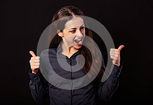 Beautiful excited shouting smiling business woman showing the fingers thumb up, the ok sign in blue shirt on black background.