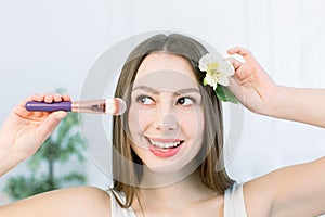 Beautiful face of young adult woman with clean fresh skin holding makeup brush in one hand and the flower in another