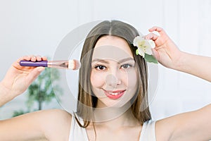 Beautiful face of young adult woman with clean fresh skin holding makeup brush in one hand and the flower in another