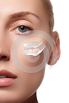 Beautiful face of young woman with cosmetic cream on a cheek. Skin care concept. Closeup portrait isolated on white. Close-up