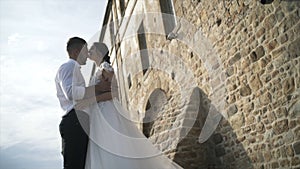 Beautiful fairytale newlywed couple posing near old castle wall. Action. The stylish groom keeping hands of the lovely