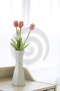 Beautiful fake pink tulips in white vase on wooden table in bed room with soft sun light and white curtains background