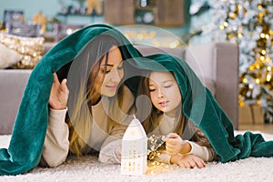 Beautiful family mom and daughter are covered with a blanket and look at the house with lights under the Christmas tree
