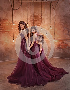 A beautiful family outfit. Mom and two daughters stand in luxurious purple-burgundy dresses with a long train against