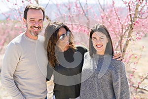 Beautiful family of three smiling cheerful and hugging on peach garden with pink petals enjoying sunny day of spring