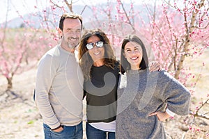 Beautiful family of three smiling cheerful and hugging on peach garden with pink petals enjoying sunny day of spring