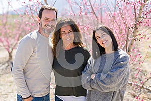 Beautiful family of three smiling cheerful and hugging on peach garden with pink petals enjoying sunny day of spring