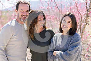 Beautiful family of three smiling cheerful and hugging on peach garden with pink petals enjoying sunny day of spring