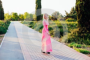 Beautiful fashion blonde woman in pink dress walks in the park at sunset