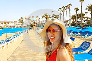 Beautiful fashion girl smiling on the beach with umbrellas, deck chairs and palm trees on the background.