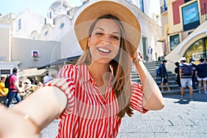 Beautiful fashion girl with striped dress and hat taking selfie photo in Capri Piazzetta square, Capri Island, Italy