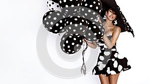 Beautiful fashion model, dressed in pinup style black dress in white polka dot and elegant hat is holding balloons with dots on