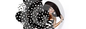 Beautiful fashion model, dressed in pinup style black dress in white polka dot and elegant hat is holding balloons with dots on