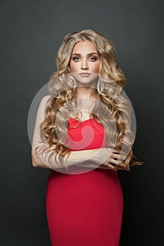 Beautiful fashion model woman with long blonde curly hair wearing red evening gown on black background