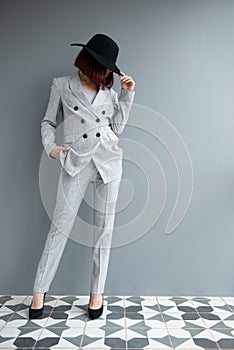 Beautiful fashion woman. Full length portrait of beautiful girl wearing light gray suit and black hat posing indoor