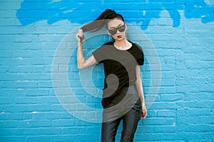 Beautiful fashionable asian girl wearing sunglasses and black clothes outfit posing in front of blue wall.