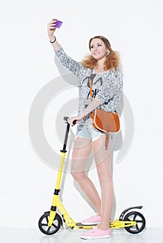 Beautiful fashionable blonde girl taking a crazy fun selfie with a vintage bag on his shoulder. Standing on a yellow scooter.