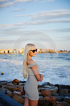 Beautiful and fashionable blonde model girl with body in the trendy striped short dress and in stylish sunglasses