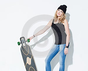 Beautiful fashionable bright young girl in jeans, a black T-shirt and hat posing with a cool longboard in the hands of near a whit