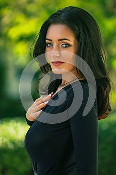 beautiful fashionable brunette woman adult posing in the park on the plants backgraund in black dress with a cleavage