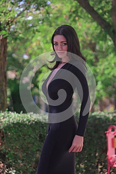 beautiful fashionable brunette woman adult posing in the park on the plants backgraund in black dress with a cleavage