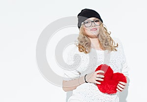 Beautiful fashionable cheerful young girl in white sweater holding a big red heart and posing near the white wall. in the studio