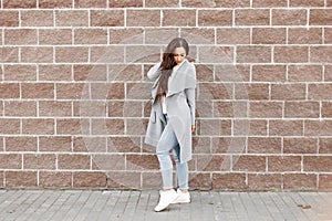 Beautiful fashionable girl in a gray coat and jeans near a brick
