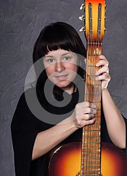 Beautiful fashionable lady wearing a gothic black dress with high collar, poses with an acoustic guitar on a grey