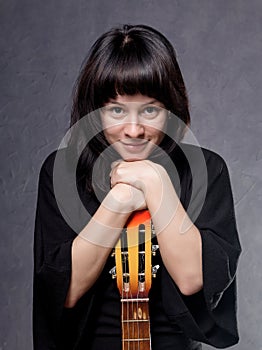 Beautiful fashionable lady wearing a gothic black dress with high collar, poses with an acoustic guitar on a grey