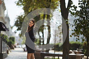 Beautiful and fashionable model girl with a shiny smile in a stylish black short dress walking by the summer city street