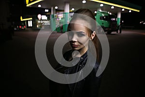 A beautiful fashionable red-haired girl poses at night on the street in the city, in autumn near a gas station