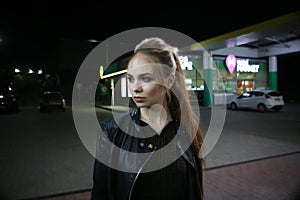 A beautiful fashionable red-haired girl poses at night on the street in the city, in autumn near a gas station