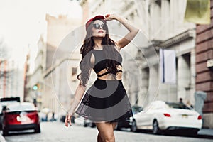 Beautiful fashionable sexy woman walking on city street wearing a black skirt, hat and sunglasses.