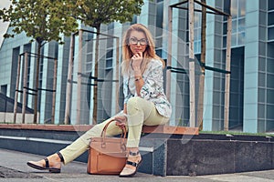 Beautiful fashionable woman in stylish clothes and glasses with a handbag, sitting on a bench against a skyscraper.