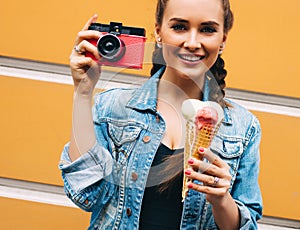 Beautiful fashionable young girl posing in a summer dress and denim jacket with pink vintage camera and multi-colored ice cream.