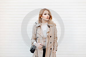 Beautiful fashionable young woman in a fashionable spring coat in a white T-shirt with a trendy black leather bag stands