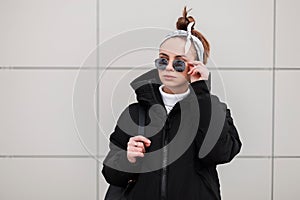 Beautiful fashionable young woman hipster with a stylish hairstyle in black glasses in a stylish black coat with a bandana with a