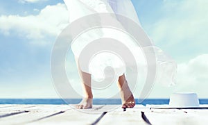 Beautiful feet of a woman in white dress on a wooden pier. Sea and sky background.