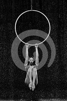 Beautiful female acrobat hanging on aerial hoop under rain on black background