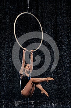 Beautiful female acrobat hanging with graceful pose on aerial hoop under rain on black background