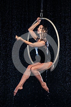 Beautiful female acrobat sitting in aerial hoop under rain on black background