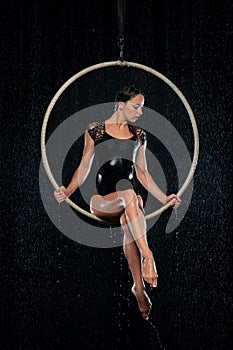 Beautiful female acrobat sitting in aerial hoop under rain on black background