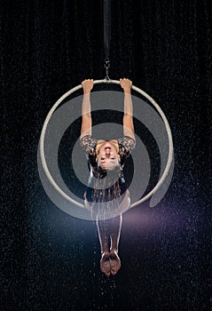 Beautiful female acrobat sitting on symmetric pose in aerial hoop under rain on black background.