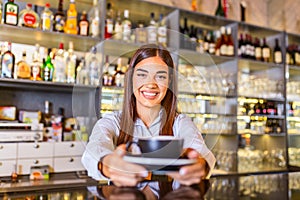Beautiful female barista is holding a cup with hot coffee, looking at camera and smiling while standing near the bar counter in