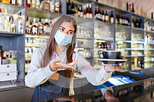 Beautiful female barista is holding a cup with hot coffee, looking at camera and wearing protective face mask while standing near