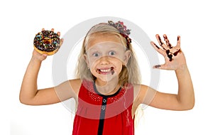 Beautiful female child with blue eyes in cute red dress eating chocolate donut with syrup stains