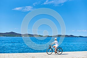 Woman riding a bicycle along stony sidewalk on blue sparkling sea water