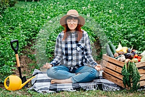 Beautiful female farmer meditating while sitting on a blanket near fresh organic vegetables in a wooden box against the background