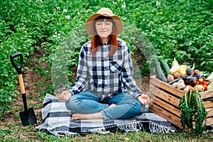 Beautiful female farmer meditating while sitting on a blanket near fresh organic vegetables in a wooden box against the background